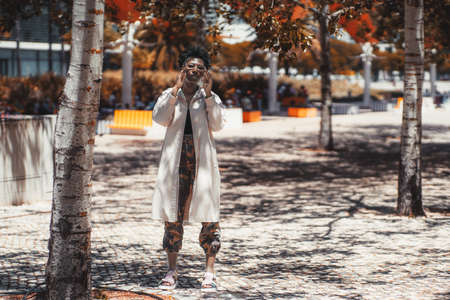 A stylish young black female in a white trench and camouflage pants, with curly Afro hair is...
