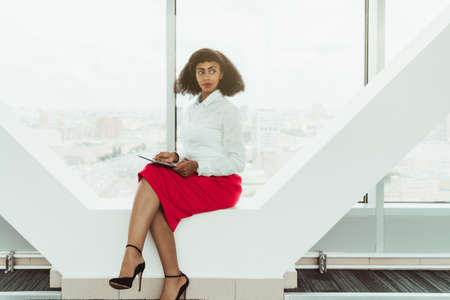 Young ravishing slender African-American businesswoman is sitting on a contemporary window bench of the modern office with a digital tablet in hands and an urban cityscape in a defocused backgroundの写真素材