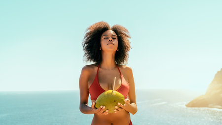 Dazzling young African-American female with curly Afro hair and coconut with a straw in her hands is raising her head up enjoying the sunlight while standing on the cliff of a seaside resortの写真素材