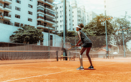 A young svelte African-American female plays tennis outdoors in the training court; a beautiful mixed woman in sports stance ready to deflect tennis ball with the racket in her hands on a sunny dayの写真素材