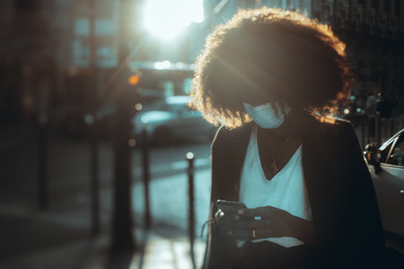 A silhouette of an African female woman in a virus protective mask phoning on a city street on a sunny evening; a black female with a curly afro hair in an anti-virus mask using a smartphone outdoorsの写真素材