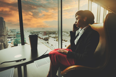 A gorgeous young African-American woman entrepreneur is talking on the phone while sitting on an armchair in front of a window of a business office skyscraper with an urban landscape and road outsideの写真素材