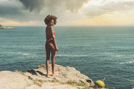 A young drop-dead gorgeous African-American woman in a swimming suit is standing half-turned on a...
