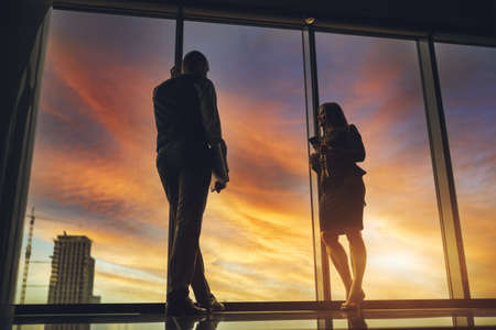 Silhouettes of two business partners near the window of a skyscraper with a stunning sunset outside and selective focus on a man entrepreneur with a laptop, his female colleague aloof using cellphoneの写真素材