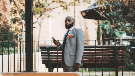A portrait of an elegant bald brutal black guy in a fashionable plaid costume and with a well-groomed beard, he is standing in the park and using his cellphone near a wooden benchの写真素材