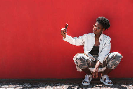 Portrait of a stylish young black female with curly Afro hair, in spectacles, white trench, camouflage trousers, taking a selfie via cellphone while squatting near a solid red wall; a copy space areaの写真素材
