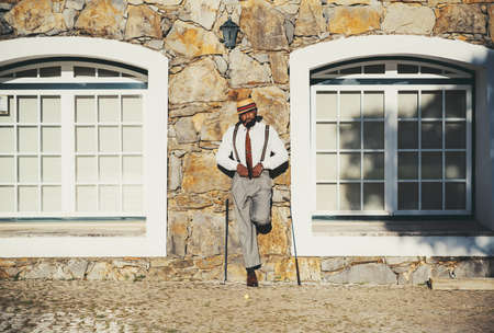 An elegant black guy in a straw hat and a fashionable outfit, with a cigar in mouth, is leaning...