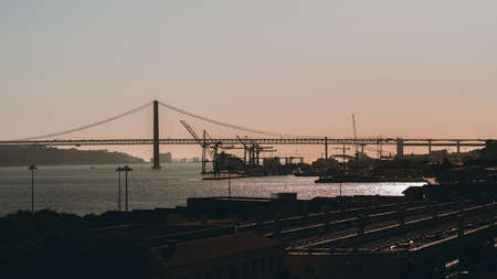 Sunset in Lisbon, Portugal: silhouettes of the Bridge 25 de Abril and docks in the background, roofs of houses, a train station in the foreground.の写真素材
