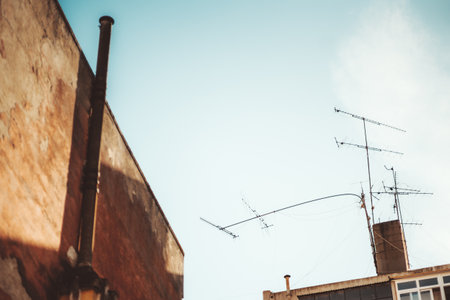 Bottom view with a shallow depth of field of a part of the wall of an old residential house with a chimney, and a roof of another one with a group of television antennas, one of which bent in the windの写真素材