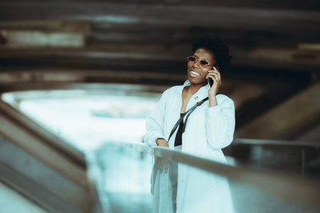 A cheerful elegant black woman in a white trench and sunglasses is laughing and having a phone conversation while standing in a modern tunnel of a railway station depot or an overhead passageの写真素材