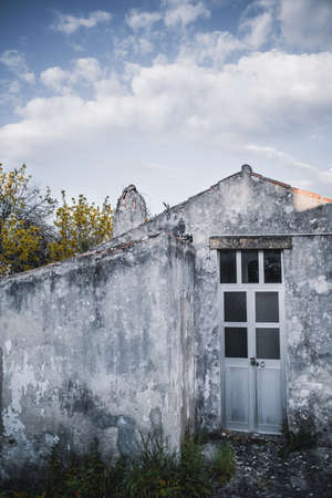 A vertical shot of a doorway of an old antique gray stone house with a triangle roof, mossy flaked walls, and narrow metal door with glass inserts; a beautiful cloudscape on the sky, Sintra, Portugalの写真素材