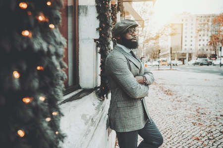Portrait of a dapper bearded handsome African guy in a woolen cap, stylish eyeglasses, and elegant custom-made business costume, leaning against the wall decorated with Christmas trees and garlandsの写真素材