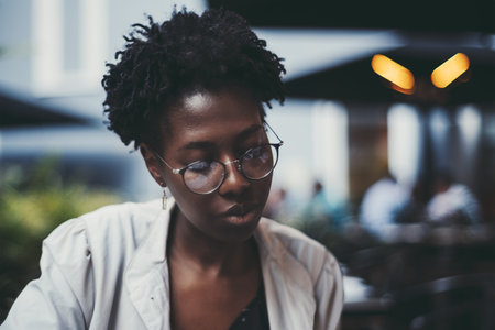 A portrait of a young black woman with curly hair sitting in a street cafe; a charming African female in spectacles and white trench is reading a menu while sitting at the table of evening restaurantの写真素材