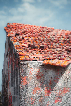 Vertical closeup view of an unfinished desolate house with a triangle tiled roof covered with reddish clay partly disordered tiles and brick wall with a thin layer of cement over it, sunny dayの写真素材