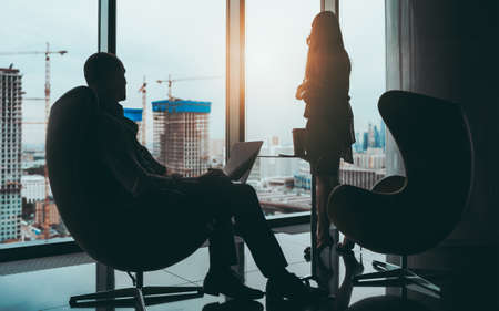 A business scene: silhouettes of two entrepreneurs near a panoramic window of a skyscraper with a construction site outside, selective focus on a businesswoman, a businessman with a laptop near herの写真素材