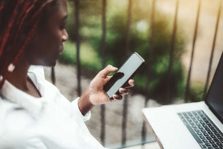 View with a shallow depth of field and selective focus on the smartphone in a hand of a young black female freelancerÂ with braided hair, sitting on the balcony with her laptop in front of herの写真素材
