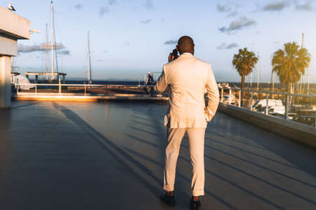 View from behind of a black guy in an elegant suit speaking on the phone while standing on a sunset coastal area; African bald man in a white costume taking on the phone outdoors, rear viewの写真素材