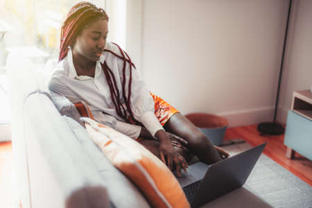 Portrait of a ravishing young black female with braided red afro hair, sitting on the couch in a cozy interior of her living room and watching a series via her laptop; a copy space place on the rightの写真素材