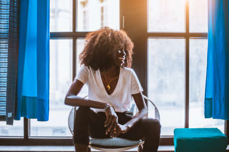 A portrait of a laughing young black female in a white t-shirt with a pendant in the shape of Africa, sitting on a chair next to the window with partly opened blue curtains, looking aside and laughingの写真素材