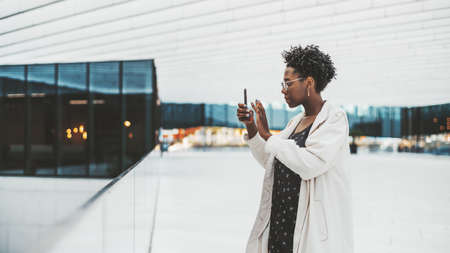 Side view of a young beautiful elegant black female in glasses, white trench coat, and with nail art, taking pics in a gallery of a modern business office building using a camera of her cellphoneの写真素材