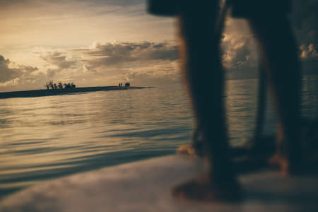 A stunning sunset seascape with shallow depth of field and selective focus on a group of people on a resort island in the background; feet of a man steering a boat in a defocused foreground, Maldivesの写真素材