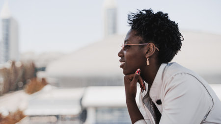 A profile portrait of a young cheerful black female with curly afro hair, in sunglasses, and a white trench in, sitting in front of a defocused cityscape background and rests her chin on her handの写真素材