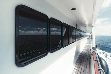 Wide-angle view of the side-deck of a luxurious white safari yacht with a row of windows of the deckhouse wooden floor of the ways and the waterscape astern with a blue ocean waterの写真素材
