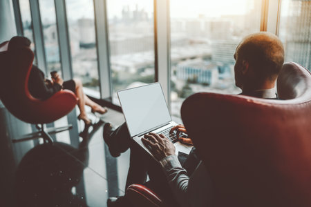 View from behind of a man entrepreneur working on his laptop, his female business partner is sitting aloof in a red armchair, with a paper cup in her hand and enjoying the view from the windowの写真素材
