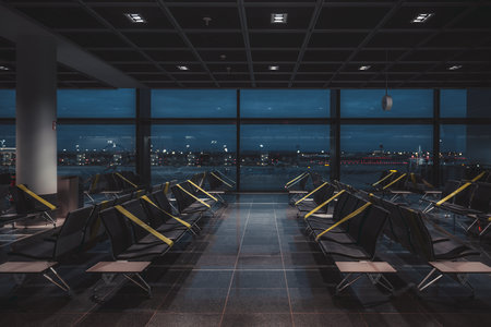 A wide-angle view of a dark empty abandoned quarantined waiting hall of a modern airport terminal at night, on a lockdown with regular greenish tapes over the seats to maintain social distancingの写真素材