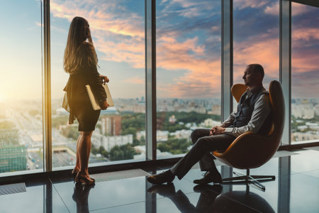 A business scene in a luxury office skyscraper near the window: a man entrepreneur pensively relaxing on an orange armchair, a businesswoman with a laptop observing the urban landscape and the sunsetの写真素材