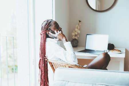 A woman sitting on a chair at the table and holding her smartphone. A young black female with braided hair wearing a white shirt, sitting and talking on the phone or listening to a voice messageの写真素材