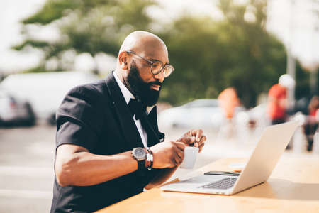 Selective focus on a stylish bearded gentleman sitting outdoors using a notebook and holding a cup....