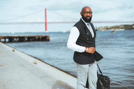 A businessman holding a messenger bag near a river, a red suspension bridge in a blurred background. A bald bearded black guy in a suit standing on the pier looking away, a copy space area on the leftの写真素材