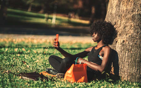 A side view of a beautiful curly-haired African female sitting on the lawn and leaning against a park tree while having a video call with her friend via a smartphone; a young black woman making selfieの写真素材