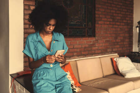 A portrait of a dazzling Brazilian female in a teal overalls sitting on the side of the sofa indoors at home and reading incoming e-mail on the screen of her phone; a copy space place on the rightの写真素材