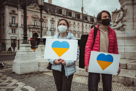 Lisbon, Portugal - February 24, 2022: Two women wearing masks and carrying placards with yellow and blue hearts in the colors of the Ukrainian flag at a protest against Russian military aggressionのeditorial素材