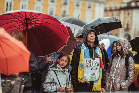 Lisbon, Portugal - March 20, 2022: a protest action in support of Ukraine, against Russian invasion and the war: row of protestants on the rainy square, a man with "Putin Killer" poster; umbrellasのeditorial素材