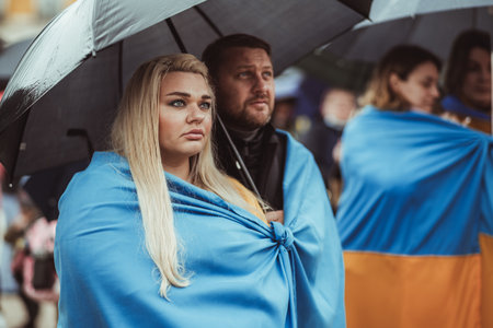 Lisbon, Portugal - March 20, 2022: a protest action in support of Ukraine, against Russian invasion and aggressive war: a couple of protesters warped in Ukrainian flag with umbrella, looking at stageのeditorial素材
