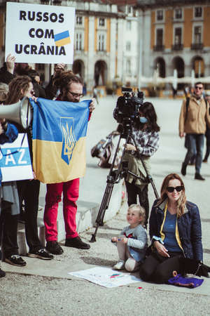 Lisbon, Portugal - March 13, 2022: protest rally against Russian incursion into Ukraine: a small girl is drawing something on the ground next to protesters with banners and flags; a TV operator behindのeditorial素材