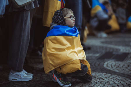 Lisbon, Portugal - April 20, 2022: protest action dedicated to saving the Ukrainian city of Mariupol from the Russian aggression; a kid in glasses wrapped in flag squats next to other protestersのeditorial素材