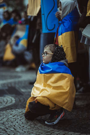 Lisbon, Portugal - April 20, 2022: protest action dedicated to saving the Ukrainian city of Mariupol from the Russian aggression; a kid in glasses wrapped in flag squats next to other protestersのeditorial素材