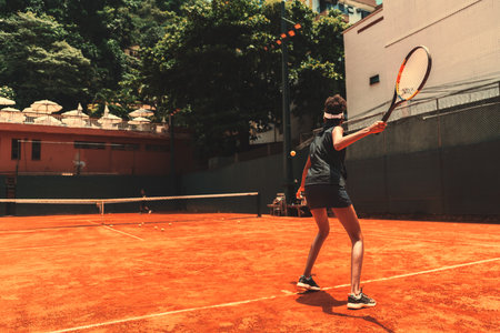 View from behind of a slim fast sportswoman on the clay field of a private sports club during her sports training session of tennis: she is swinging her racquet to hit back a yellow tennis ballの写真素材