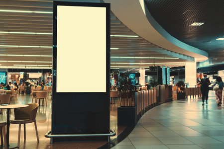 An empty vertical advertising banner mockup in front of a crowded shopping mall or an airport food court with tables and customers in the background and a copy space place on the right for an ad textの写真素材