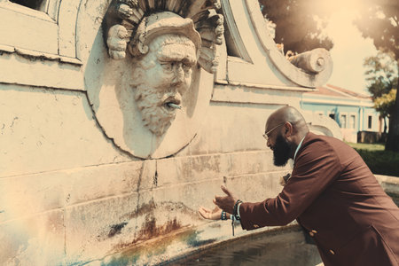A side view of a mature bald black man with a beard, in a tailored suit of a chestnut color and eyeglasses, washing his hands in the water of a street antique marble fountain or going to drink from itの写真素材