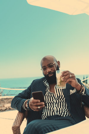 A vertical shot of a handsome elegant mature bald African guy with a black beard is drinking a caipirinha cocktail and fiddling with his cell phone while sitting in a coastal cafe on a warm sunny dayの写真素材