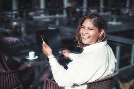 A portrait of a dazzling laughing Hispanic plus-size businesswoman sitting in an outdoor cafe with her laptop and a cup of coffee and holding a smartphone in her hand; a Brazilian woman with cellphoneの写真素材