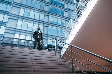 An elegant African man entrepreneur is standing on the stairs of a yard of a business skyscraper; a black businessmanÂ in a fashionable outfit with a bag on the stairway of an office high-riseの写真素材