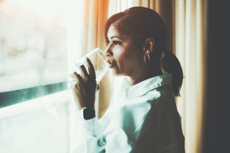 A side view with selective focus on an adult beautiful plus-size Hispanic female with a smartwatch drinking the wine from the glass and pensively looking outside the windowの写真素材