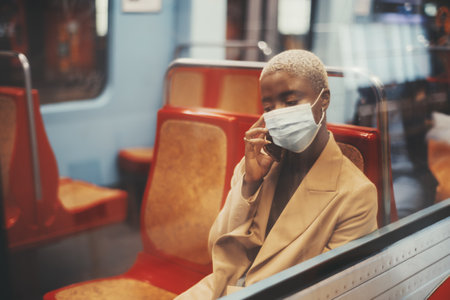 A portrait with a shallow depth of field of a young black lady with short hair painted in white color, with a mask on her face, sitting alone on a red seat of a subway train and talking on the phoneの写真素材
