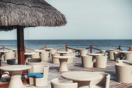 A large group of wicker tables and armchairs on a wooden floor of an outdoor cafe or a restaurant of a luxury tropical resort with a part of a straw canopy roof; an ocean horizon in the backgroundの写真素材